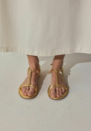 Kājām zelta krāsas sandales ar siksniņām un gliemežvākveida rotājumiem, tērptām garā bēša krāsas svārkā, stāvot uz gaiša grīdas seguma.