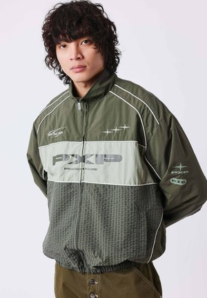 Homme aux cheveux bouclés portant une veste zippée verte et grise avec des passepoils blancs et des logos sur le devant et la manche, debout sur un fond uni.