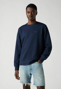 Marineblauwe sweatshirt met een ronde hals en gebreide manchetten. Voorzien van rode geborduurde tekst op de borst. Gemaakt in combinatie met lichte denim shorts.