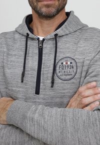 Gray zip-up hoodie with a textured finish, navy accents, and a circular logo on the chest featuring a vintage design.