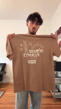Hombre con cabello rizado sosteniendo una camiseta marrón con un diseño de plantas y notas musicales y el texto "Plantas de Interior, Nutre tus Raíces, Levi's"