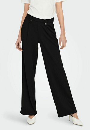 Pantalons noirs à jambes larges en tissu léger avec des plis avant et deux boutons à la taille, associés à des talons blancs pointus.