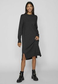 Abito in maglia grigio scuro a maniche lunghe con spacchi laterali, vestibilità rilassata, colletto e polsi a coste. Indossato con stivali neri a tacco largo.