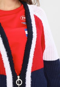 Virkat cardigan i röd, vit och marinblå med öppen front, texturerad tyg och stort silverfärgat dragkedjedetalj. Korta ärmar och randig mönster.