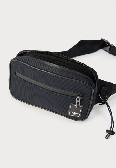 Μαύρο υφασμάτινο ορθογώνιο τσαντάκι μέσης με μπροστινή τσέπη με φερμουάρ, ρυθμιζόμενο λουράκι και μεταλλικό έμβλημα λογότυπου σε λευκό φόντο.