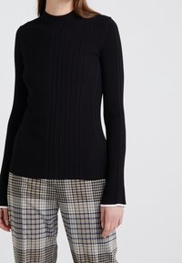 Zwarte geribbelde lange mouw top met een hoge hals, voorzien van een textuurdesign. Gecombineerd met een broek met ruitpatroon in neutrale tinten.