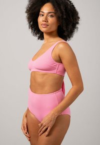 Kvinna med lockigt hår som bär en rosa högmidjad bikiniunderdel och matchande bikiintopp, stående mot en plain ljusgrå bakgrund.