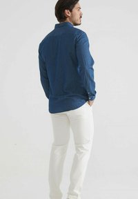 Chemise en denim bleu foncé, à manches longues, présentant un poignet et un col boutonnés. Assortie à un pantalon blanc. Tissu texturé.