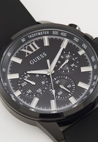 Svart chronograph klokke med teksturerte sub-dials, sølvfargede timeindekser og tall. Har tachymeter og gummirem. Merke: Guess.