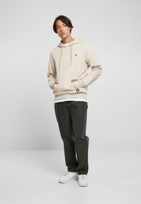 Beige, bomullsblanding hettegenser med kengurulomme, snor i hetta og logo-patch på brystet, kombinert med mørke jeans og hvite sneakers.