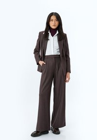 Ruskea pinstripe- käsityöläissakko ja leveälahkeiset housut, yhdistetty valkoiseen paitaan ja purppuraan poolopaitaan, neutraalia taustaa vasten.