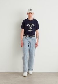 Marinblå t-skjorte med hvit trykt tekst, lyseblå avslappede jeans, hvite joggesko og en hvit caps med logo. Enkel, uformell estetikk.