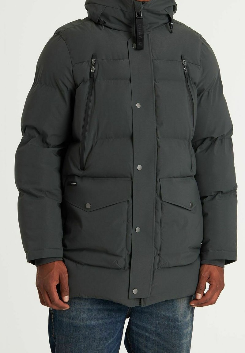 La chaqueta acolchada de color gris oscuro cuenta con un cierre frontal con cremallera, capucha ajustable y múltiples bolsillos con cierres de botón a presión. Textura suave y diseño elegante.