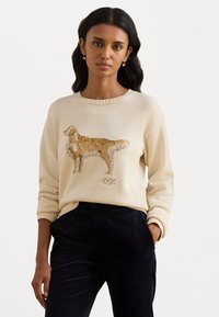 Woman with dark hair wearing cream sweater featuring a golden retriever knit design and black pants, standing against a plain white background.