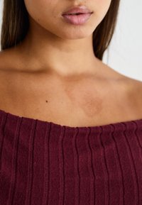 Burgundy, bordás, lecsúsztatott vállú felső, puha anyagból, textúrált felületet és finom fényt mutatva, kiemelve a dekoltázst és a kulcscsontot.