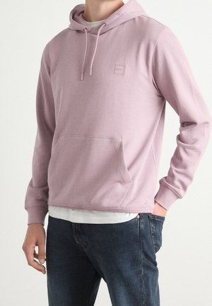 Mand iført en lyse lilla hættetrøje med frontlomme og mørkeblå jeans, hånd i lommen.