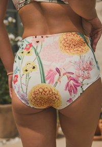 Hoog uitgesneden bikini-bovenstukje in een witte stof met levendige bloemmotieven in roze, geel en groen, voorzien van decoratieve hardware in de taille.