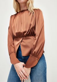 Blouse en satin marron avec corsage froncé, manches longues bouffantes et détails de poignets boutonnés ; ajustée à la taille et légèrement plus longue à l'arrière.