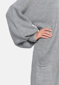 Cardigan in maglia grigio con una vestibilità rilassata, maniche oversize e una tasca sul lato sinistro. Tessuto testurizzato con un motivo a coste.