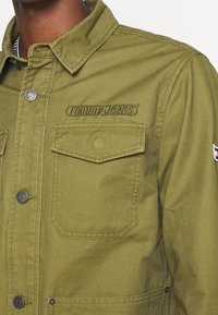 Detailní záběr na olivově zelenou bundu s klopovým hrudním kapsou, kovovými knoflíky, výšivkou "Tommy Jeans" na prsou a malou nášivkou vlajky na rukávu.