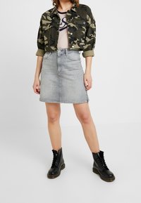 Krótkia kurtka w kamuflażu, jasnoszara spódnica dżinsowa i czarne wiązane buty. Spódnica ma prosty krój i zapięcie na guziki z przodu.