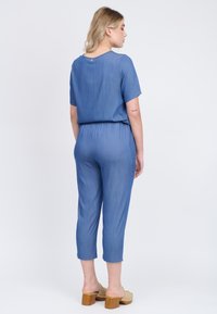 Blauer Denim-Overall mit kurzen Ärmeln und elastischem Bund, mit verkürzten Beinen und einer glatten Textur. Es werden hellbraune Clogs getragen.