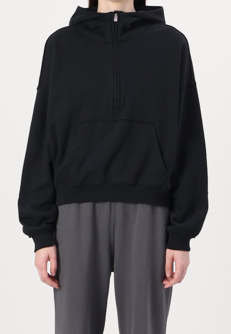 Personne portant un sweat à capuche noir avec une fermeture éclair à l'avant et une poche kangourou, associé à un pantalon gris ample.