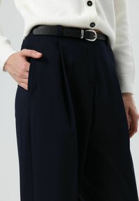 Zwarte leren riem met zilveren gesp, gedragen over marineblauwe geplooide broek. Witte gebreide cardigan met gedeeltelijk zichtbare knopen.