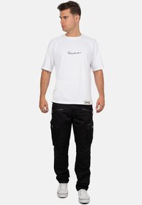 T-shirt bianco in cotone con maniche corte, con logo in nero scritto. Pantaloni cargo neri con cerniere e molte tasche, abbinati a sneakers bianche.