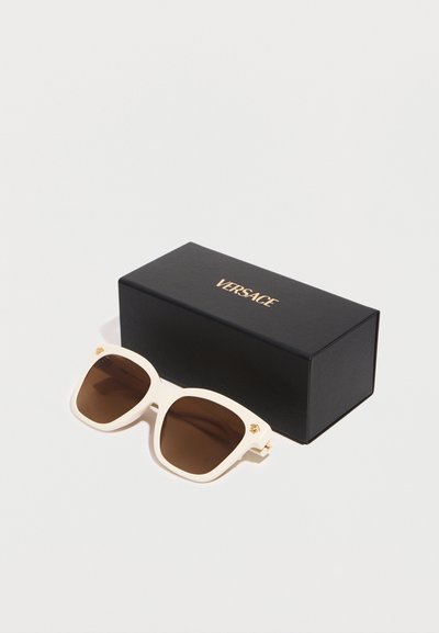 Lunettes de soleil Versace à monture blanche avec des verres marron, posées à côté d'un étui noir de la marque Versace sur un fond blanc.