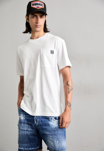 T-shirt bianco in cotone con taschino sul petto e logo "D2" nero, abbinato a pantaloni corti in denim blu strappati con dettagli di macchie di vernice.