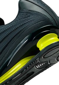 Zwarte sportschoen met een bovenwerk van getextureerd mesh, glanzende zwarte accenten en een felgeel dempingsdetail op de buitenzool.