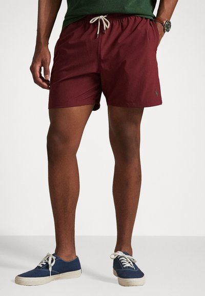 Pantaloni da bagno bordeaux con cintura elastica, dettaglio con cordoncino e piccolo logo ricamato. Indossati con scarpe in tela blu.