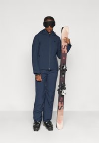 Chaqueta de esquí y pantalones de color azul oscuro, botas de esquí negras, sosteniendo un esquí rosa y marrón. Presenta un cierre frontal con cremallera y gafas para protección.