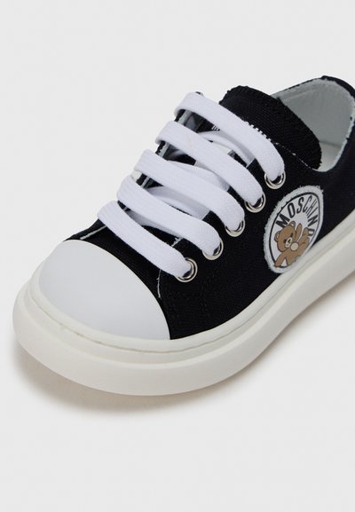 Μαύρο πάνινο αθλητικό παπούτσι με λευκή λαστιχένια μύτη και σόλα. Διαθέτει λευκά κορδόνια και στρογγυλό μπαλώμα με σχέδιο αρκούδας.