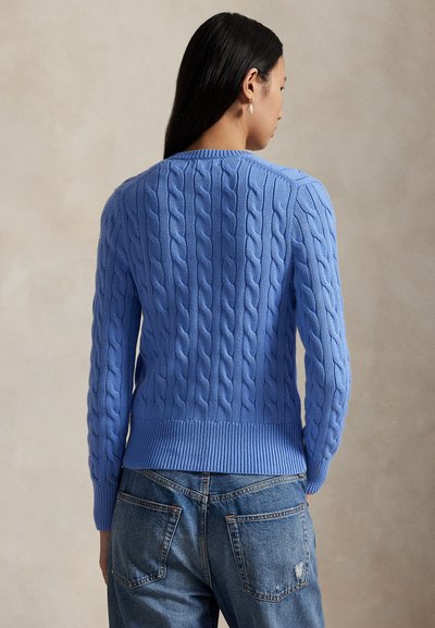 Femeie cu păr lung și închis la culoare, purtând un pulover tricotat cu model în nuanță de albastru deschis și blugi albaștri, privindu-se cu spatele spre un fundal neutru.