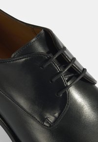 Chaussure en cuir noir à bout rond, texture lisse et détails cousus. Présente un système de laçage standard et une surface élégante et polie.