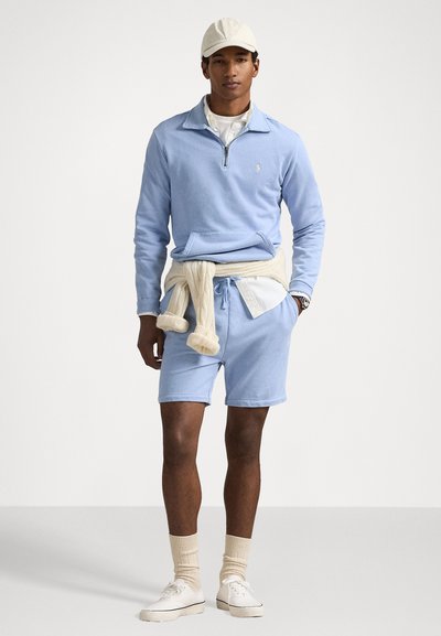 Pulover din fleece de culoare albastru deschis, cu fermoar la jumătate, pantaloni scurți asortați și șosete bej. O bluză crem este legată în jurul taliei.