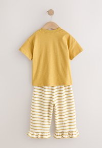 Chemise jaune à manches courtes associée à un pantalon blanc avec des rayures horizontales moutarde et des ourlets à volants, suspendue à un cintre en bois.