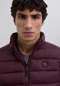 Jonge man met donker haar in een maroon gekleurde donsjack met rits en een doodskop met gekruiste botten logo op de borst, neutrale grijze achtergrond.