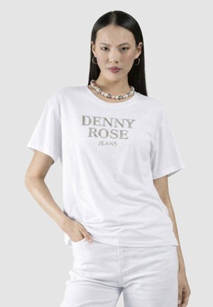 Donna con capelli lisci neri indossa una maglietta bianca "Denny Rose Jeans", pantaloni bianchi, collana di perle e orecchini a cerchio, posando con la mano in tasca.