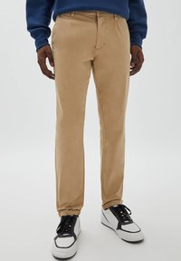 Beige, maßgeschneiderte Hose mit geradem Bein, gefertigt aus glattem Stoff. Kombiniert mit schwarz-weißen Sneakern im Low-Top-Stil.