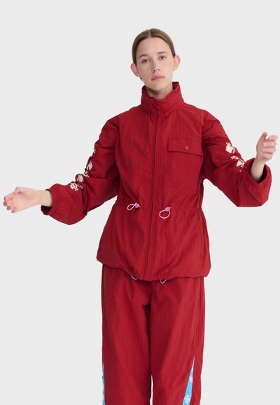 Jeune femme portant une veste rouge à fermeture éclair avec des broderies florales sur les manches et un pantalon rouge assorti à bandes latérales bleues, debout les bras légèrement levés.