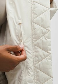 Kerged vest beežikas värvusega, millel on tikandiga muster. Omab lukuga küljetaskut ja sileda, läikelise kangasteksti.