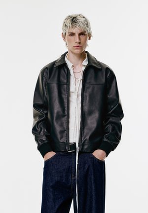 Jeune personne aux cheveux blonds courts portant une veste en cuir noir, une chemise blanche à boutons, un jean bleu, se tenant avec les mains dans les poches.