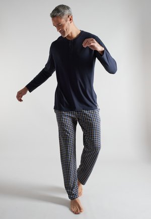 Mann mit grauen Haaren, der ein marineblaues Langarmshirt und blau-weiß-gelbe karierte Pyjamahosen trägt, steht barfuß vor einem schlichten Hintergrund.