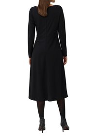 Eine Frau trägt ein langärmliges schwarzes Midikleid mit schwarzen Strumpfhosen und schwarzen, knöchelhohen Stiefeletten und steht mit dem Rücken zur Kamera.