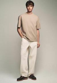 Beige Baumwoll-T-Shirt mit kurzen Ärmeln, kombiniert mit weißen weit geschnittenen Hosen und schwarzem Schuhwerk mit braunen Gummisohlen und Schnürsenkeln.