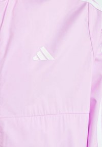 Světle růžová bunda vyrobená z hladkého materiálu, s bílým logem Adidas na přední straně. Design zahrnuje kontrastní švy a elastický pas.