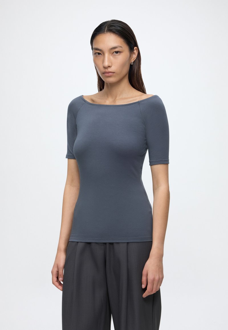Femme aux cheveux raides et foncés portant un haut ajusté à manches courtes gris foncé et un pantalon ample gris foncé sur un fond clair uni.